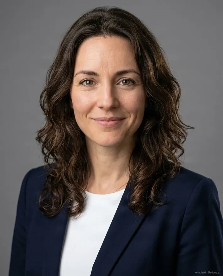 The same invented woman as a corporate headshot: a grey studio backdrop, a dark blazer, a small AI-generated mark in the corner. Not a real person.